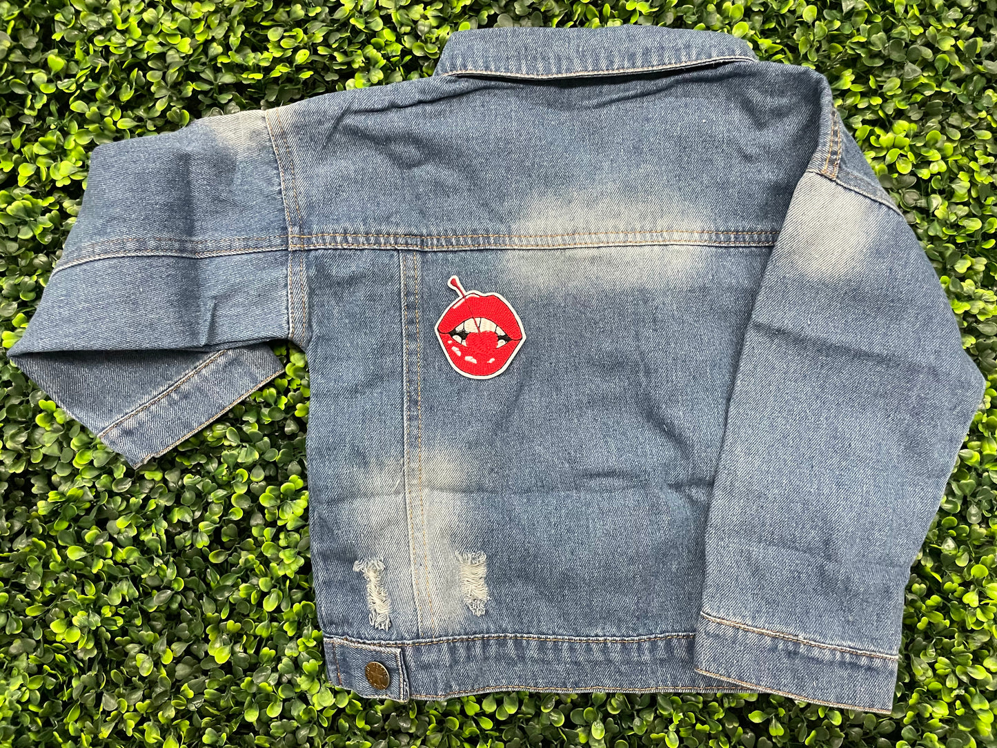 Jean jacket with  a red lips embroidered patch with a cherry, with greenery in the background.