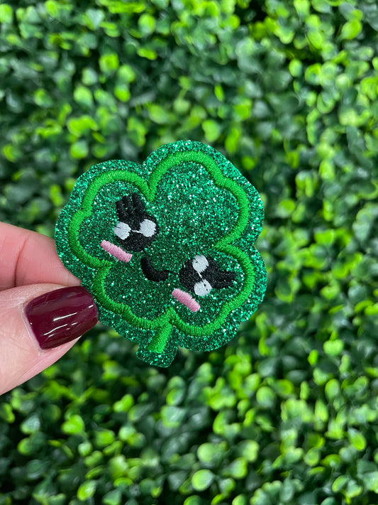 Embroidered patch of a green kawaii clover, placed on a green leafy background