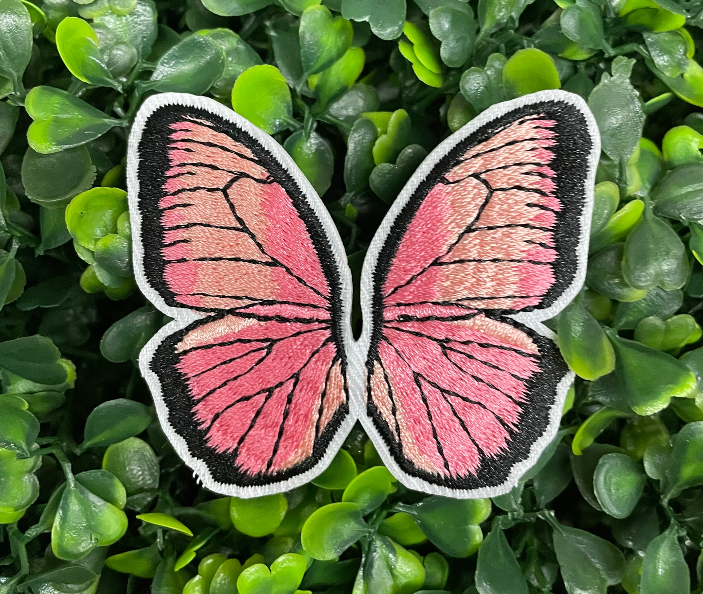 A pink butterfly embroidered patch held against a leafy background.
