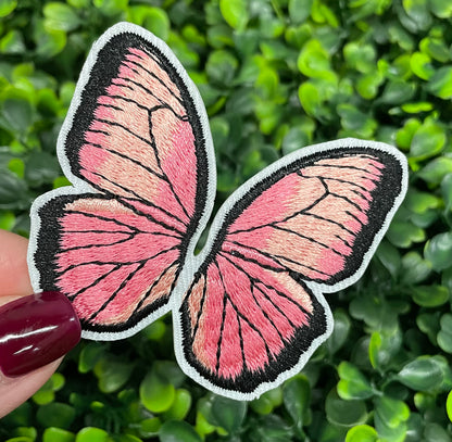A pink butterfly embroidered patch held against a leafy background.
