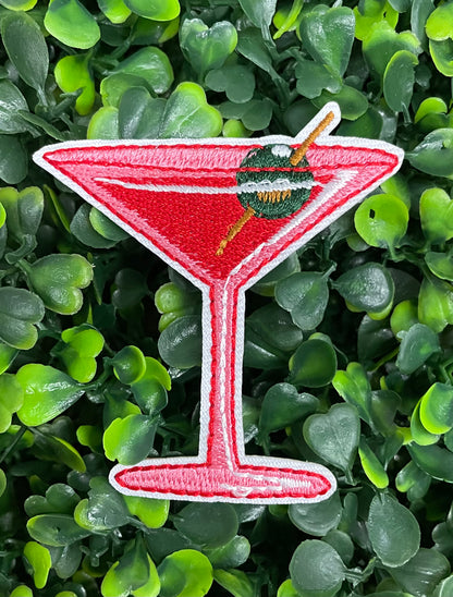 An iron-on patch depicting a red martini glass with a green olive, set against a blurred background of greenery.