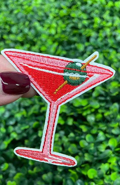 A hand holding an iron-on patch depicting a red martini glass with a green olive, set against a blurred background of greenery.