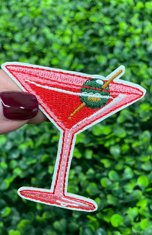 A hand holding an iron-on patch depicting a red martini glass with a green olive, set against a blurred background of greenery.