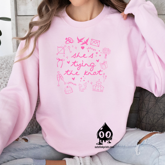 A light pink text sweatshirt with 'She's Tying the Knot' printed on the front, surrounded by various symbols and illustrations related to weddings.