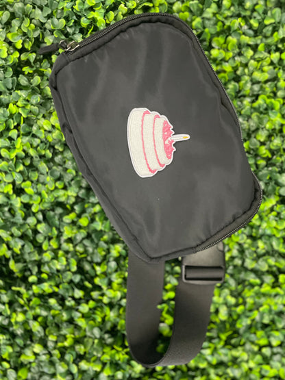 A small pink and white birthday cake embroidered patch placed on top of a small black bag