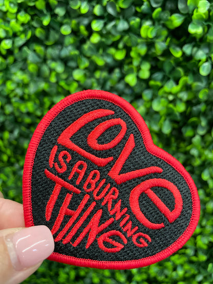 Heart-shaped patch with 'Love is a Burning Thing' text held against a green leafy background