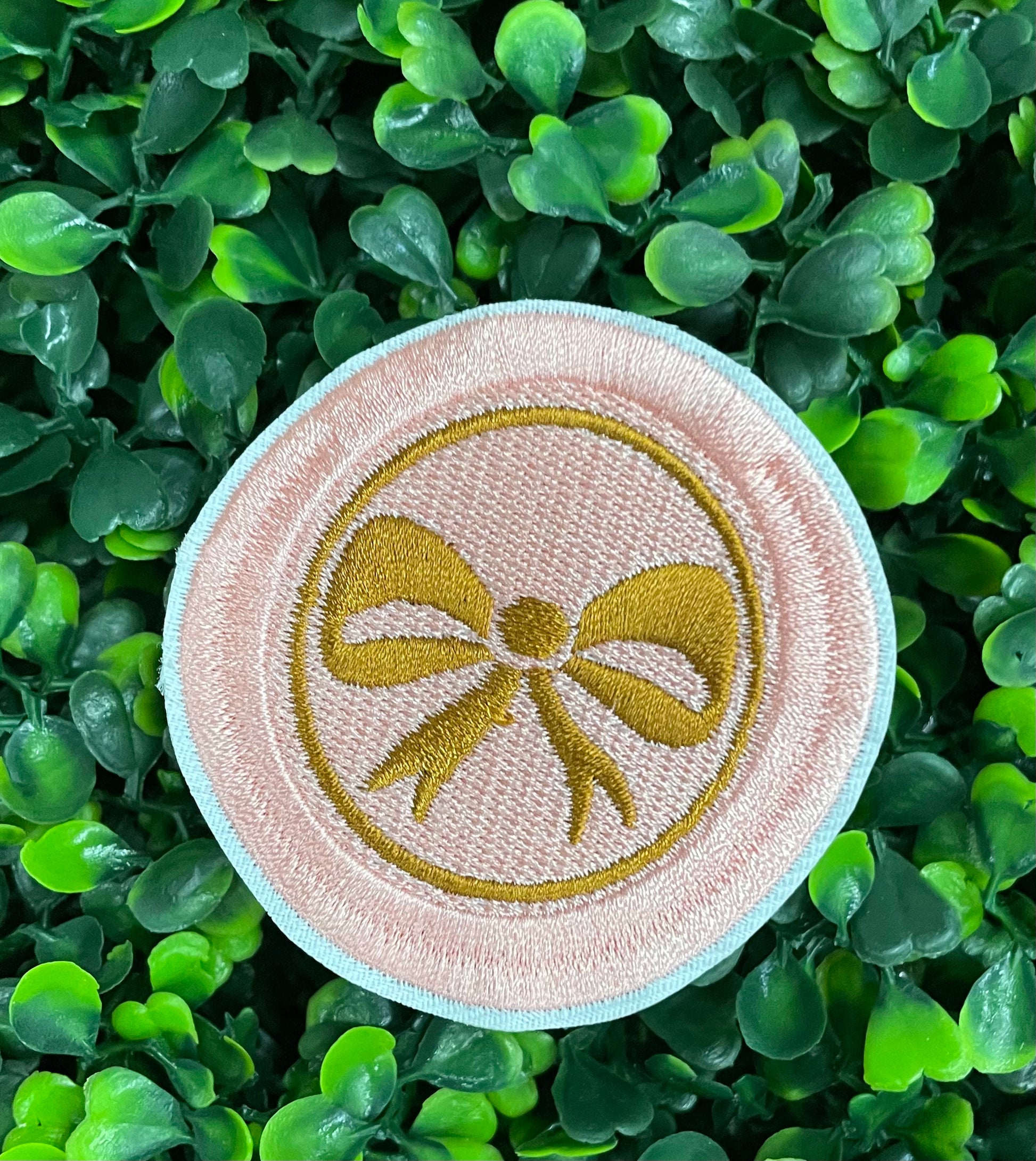 A round patch featuring a gold bow on a pink background with a embroidered edge, placed on a leafy green backdrop