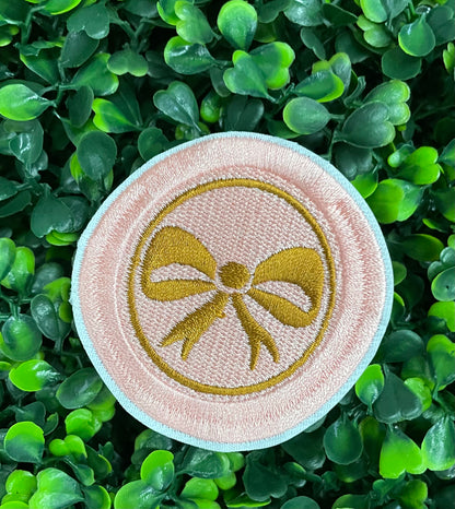 A round patch featuring a gold bow on a pink background with a embroidered edge, placed on a leafy green backdrop