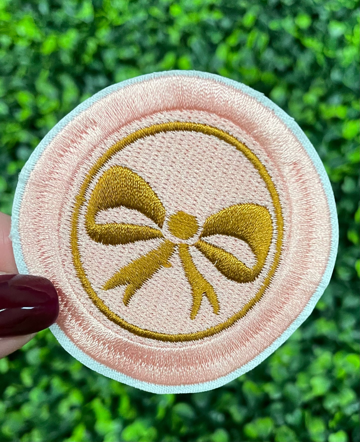 A round patch featuring a gold bow on a pink background with a embroidered edge, placed on a leafy green backdrop