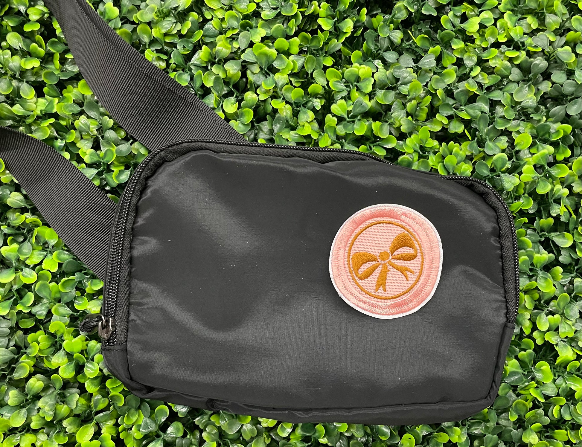 A round patch featuring a gold bow on a pink background with a embroidered edge, placed on a black shoulder bag