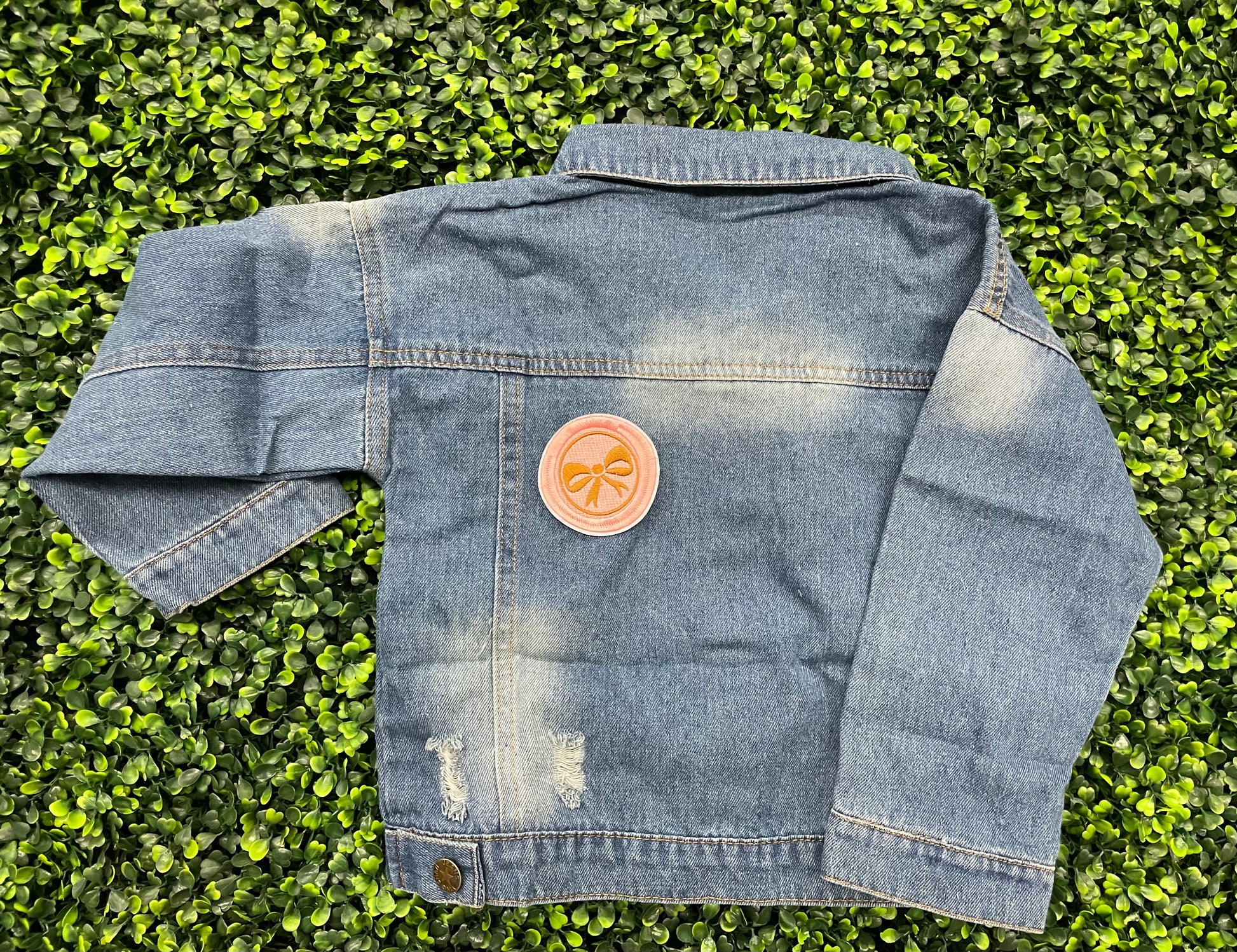 A round patch featuring a gold bow on a pink background with a embroidered edge, placed on a child's jean jacket