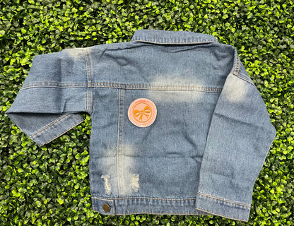 A round patch featuring a gold bow on a pink background with a embroidered edge, placed on a child's jean jacket