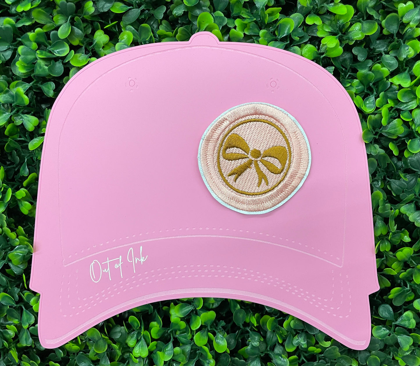 A round patch featuring a gold bow on a pink background with a embroidered edge, placed on a pink hat graphic