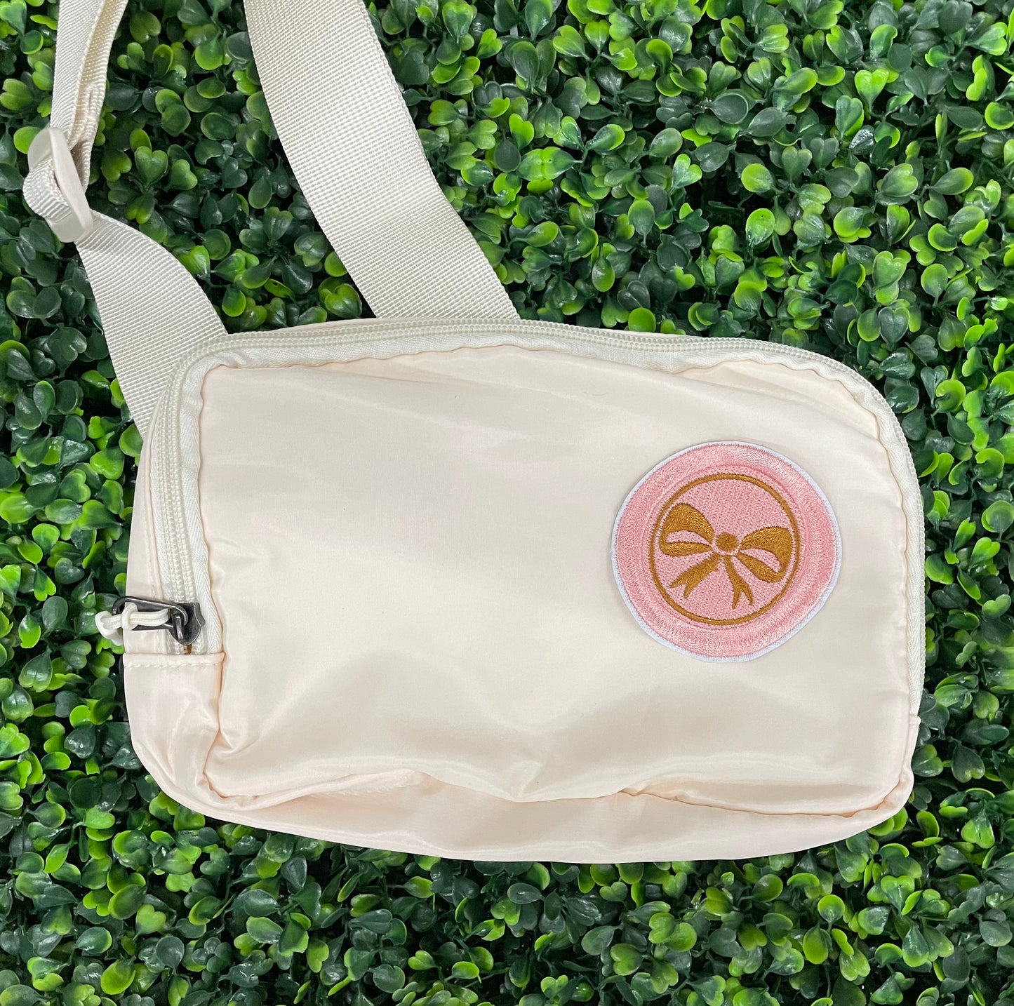 A round patch featuring a gold bow on a pink background with a embroidered edge, placed on a white shoulder bag