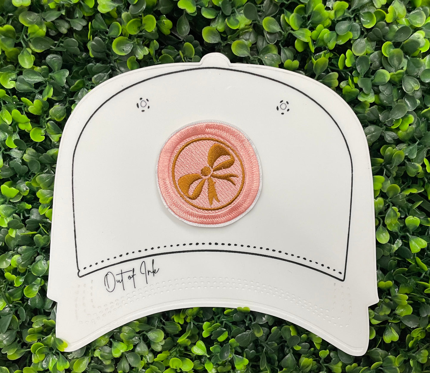 A round patch featuring a gold bow on a pink background with a embroidered edge, placed on a white hat