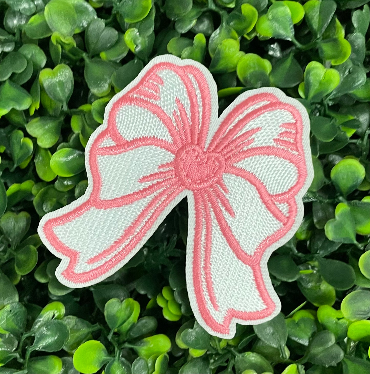 A hand holding a white and pink heart-shaped bow patch with an embroidered design, placed on a green backdrop
