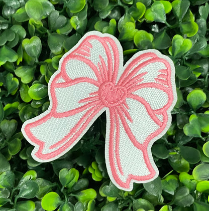 A hand holding a white and pink heart-shaped bow patch with an embroidered design, placed on a green backdrop