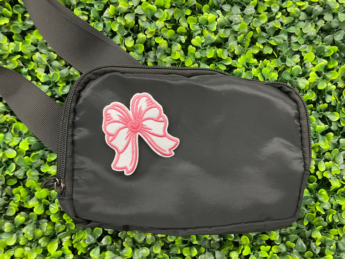 A hand holding a white and pink heart-shaped bow patch with an embroidered design, placed on a black shoulder bag