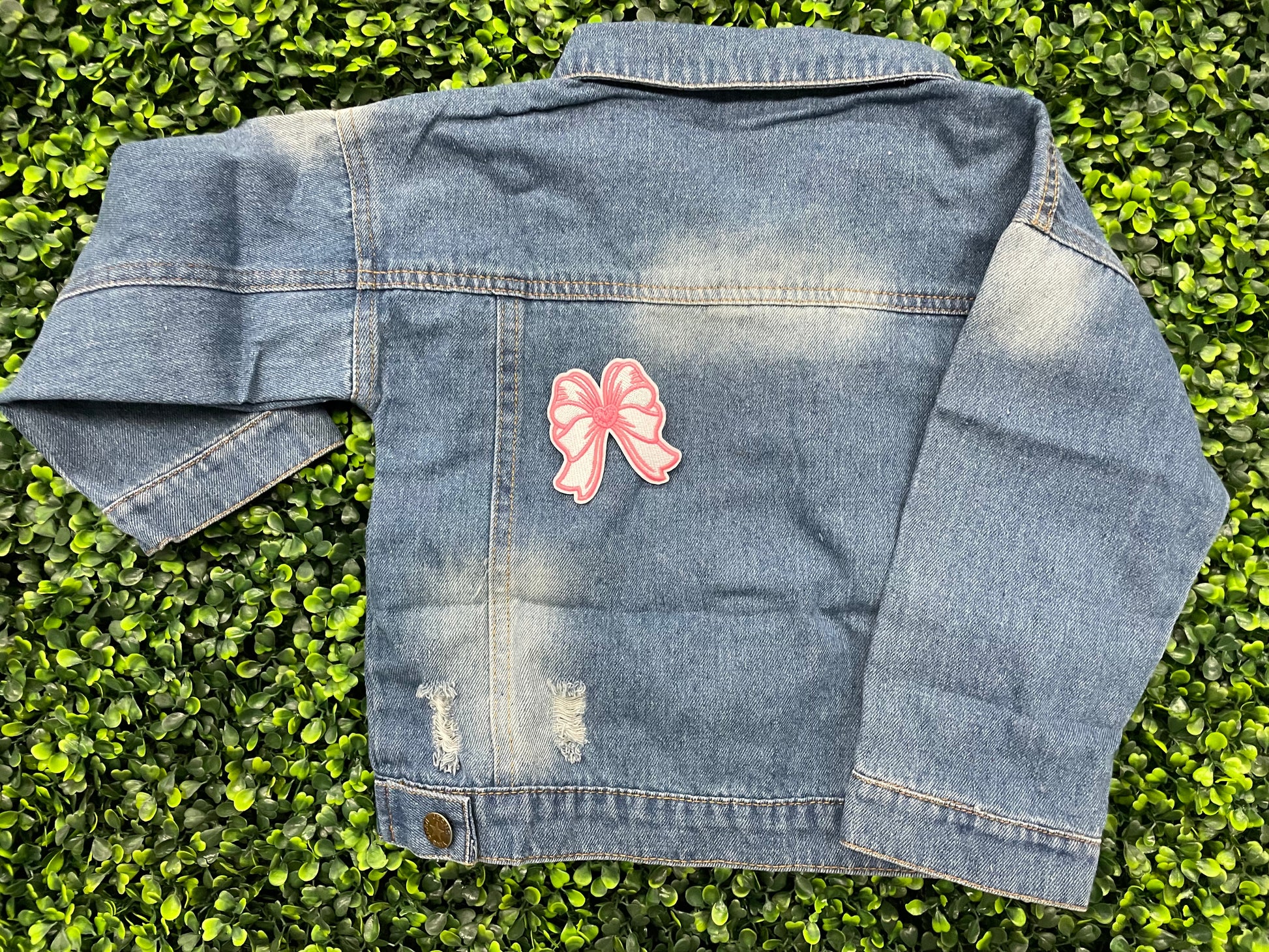 A hand holding a white and pink heart-shaped bow patch with an embroidered design, placed on a child's jean jacket