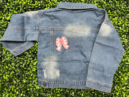 A hand holding a white and pink heart-shaped bow patch with an embroidered design, placed on a child's jean jacket