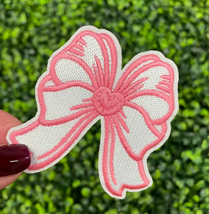 A hand holding a white and pink heart-shaped bow patch with an embroidered design, placed against a green leafy background.