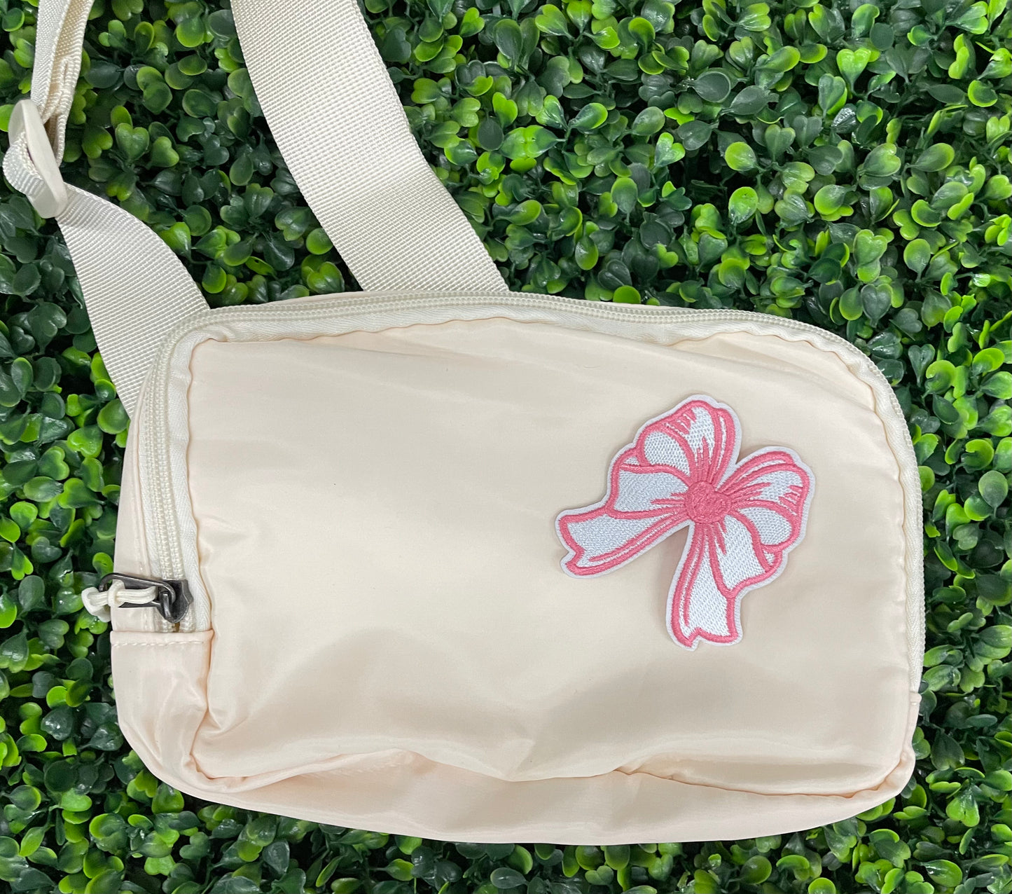 A hand holding a white and pink heart-shaped bow patch with an embroidered design, placed on a white shoulder bag against a green backdrop