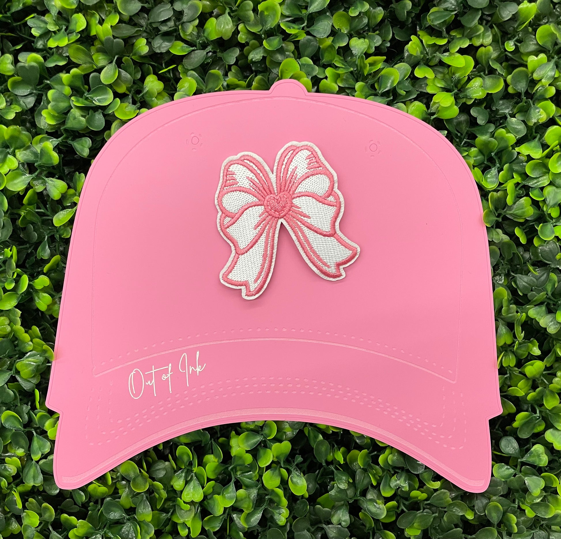 A hand holding a white and pink heart-shaped bow patch with an embroidered design, placed on a pink hat against a green backdrop