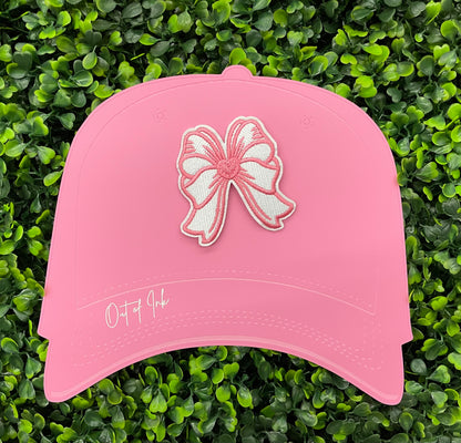 A hand holding a white and pink heart-shaped bow patch with an embroidered design, placed on a pink hat against a green backdrop