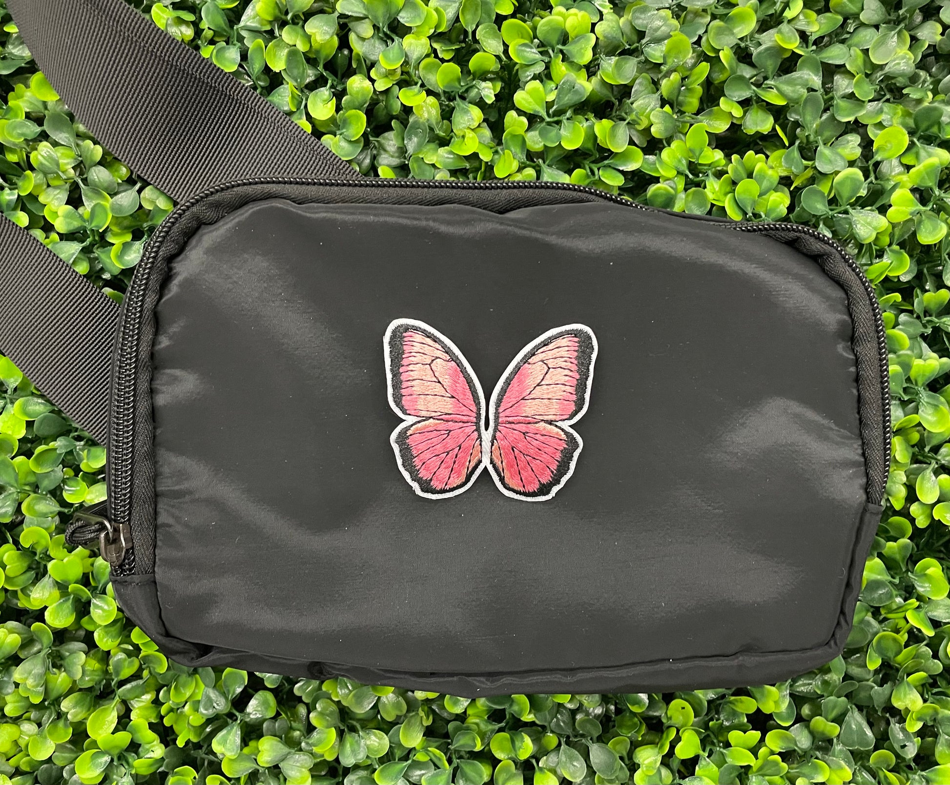 A pink butterfly embroidered patch held onto a black shoulder bag against a leafy background