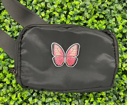 A pink butterfly embroidered patch held onto a black shoulder bag against a leafy background