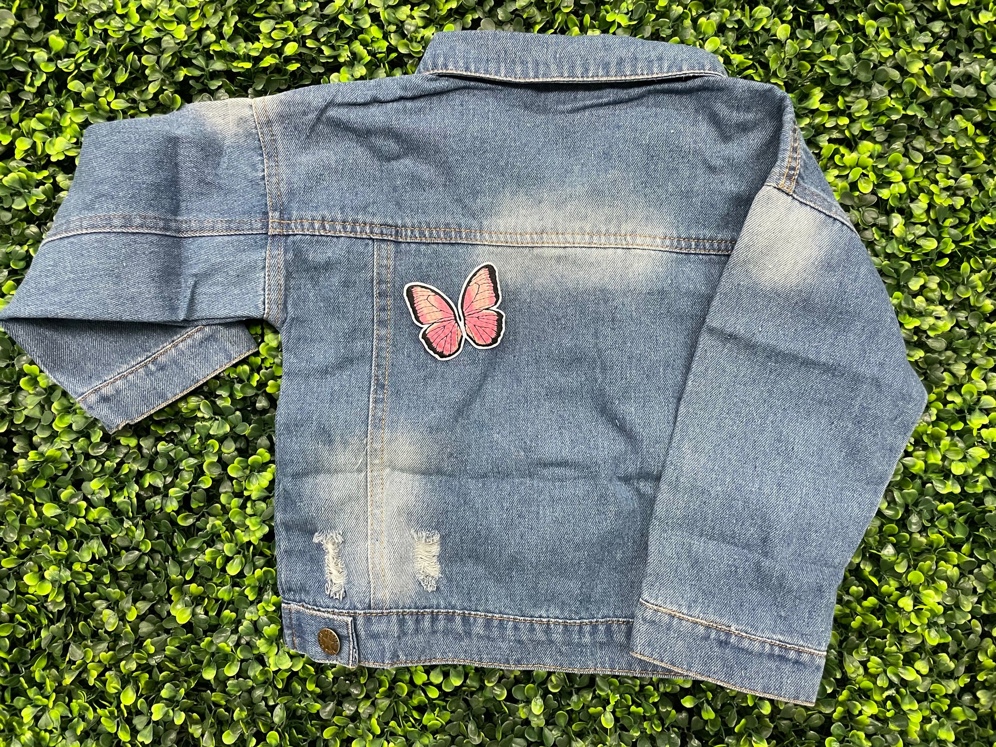 A pink butterfly embroidered patch held onto a jean jacket against a leafy green background