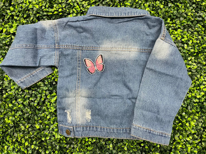 A pink butterfly embroidered patch held onto a jean jacket against a leafy green background