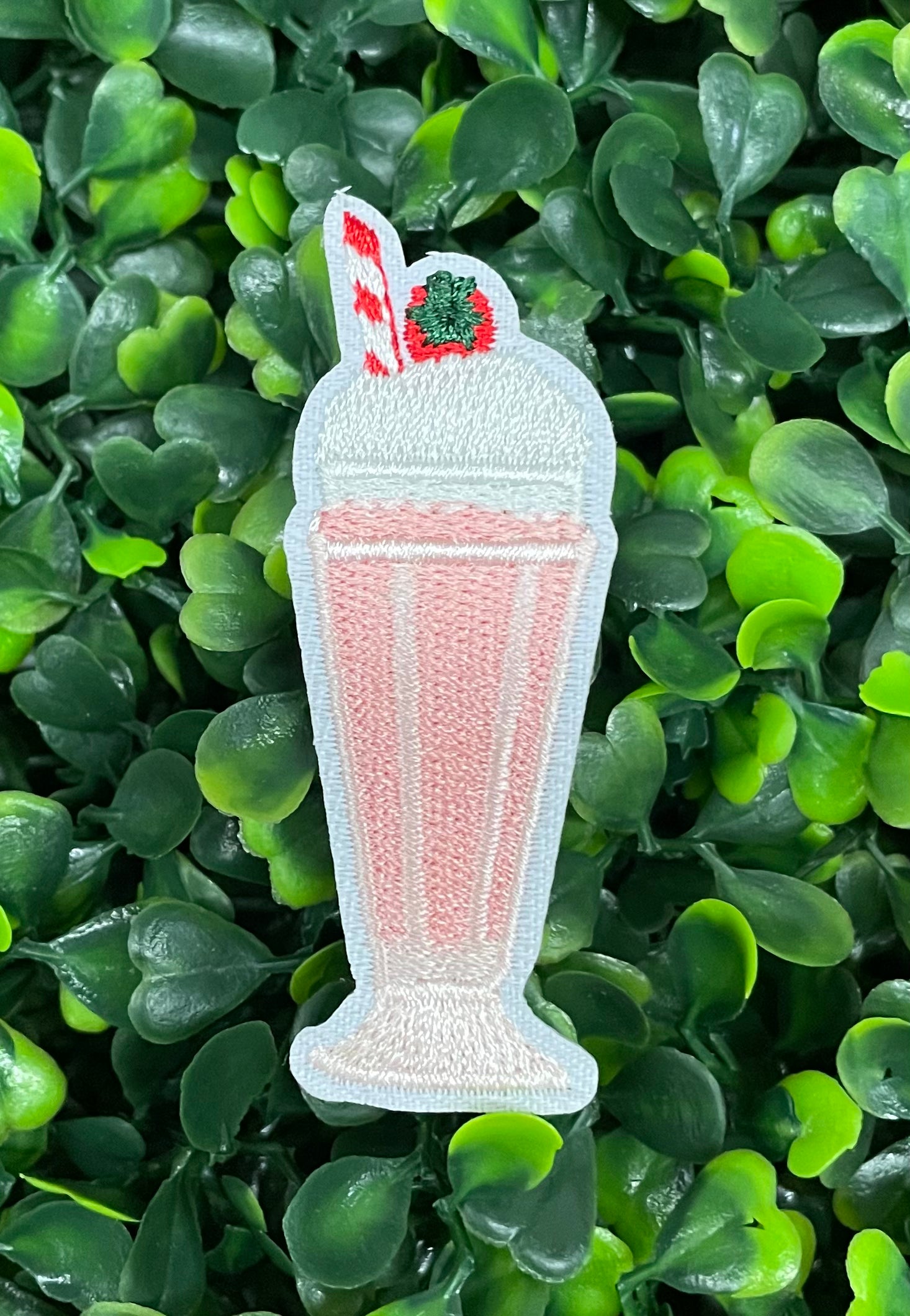 A pink milkshake embroidered patch with a cherry on top, held against a green leafy background.