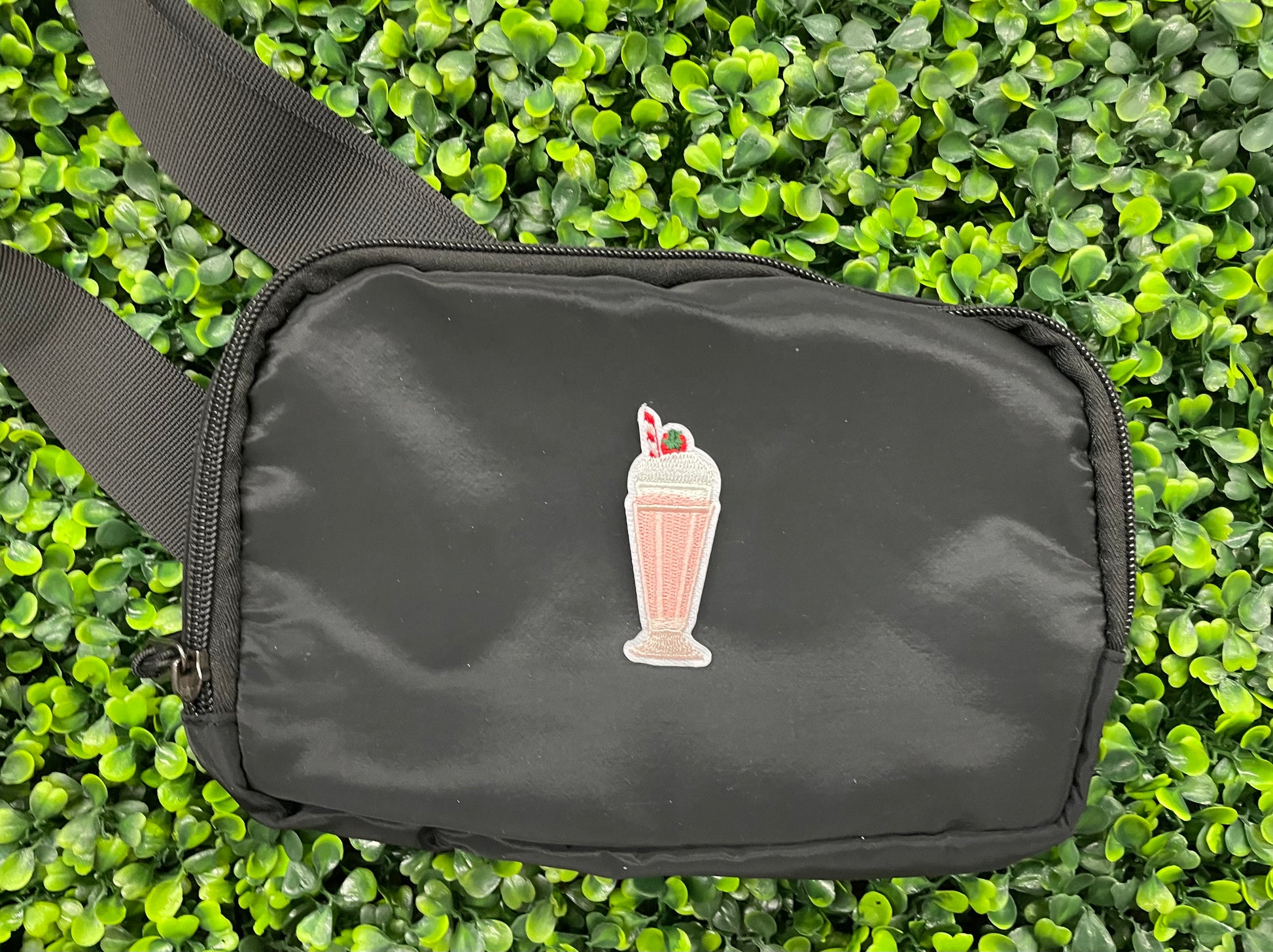 A pink milkshake embroidered patch with a cherry on top, held onto a black shoulder bag