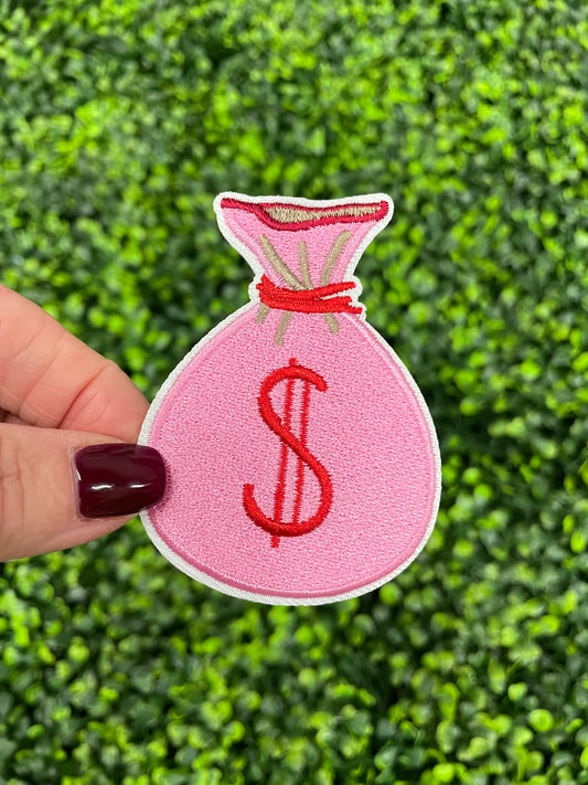 A pink embroidered patch in the shape of a money bag with a dollar sign in red, held against a green leafy background.