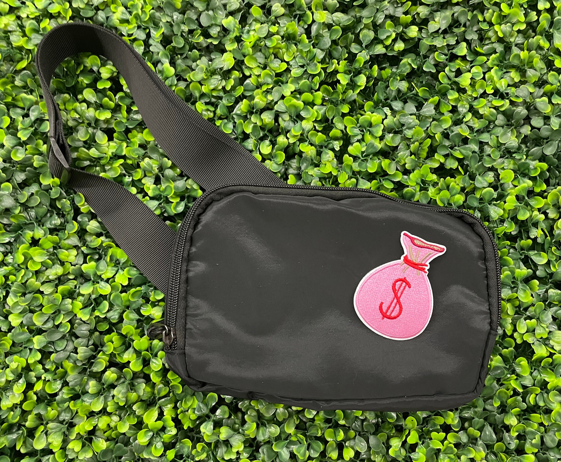 A patch depicting a pink bunched money bag with a red dollar sign, placed on a black shoulder bag
