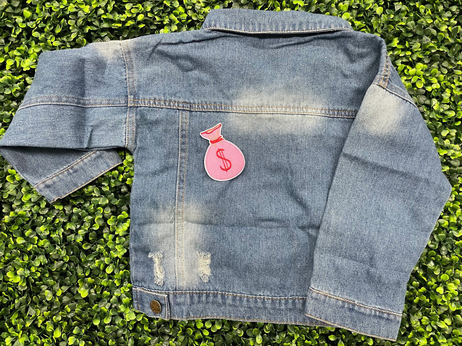 A patch depicting a pink bunched money bag with a red dollar sign, placed on a jean jacket