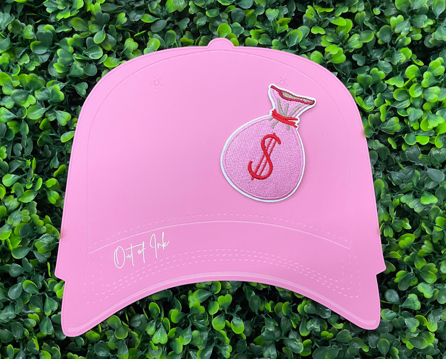 A patch depicting a pink bunched money bag with a red dollar sign, placed on a pink hat graphic