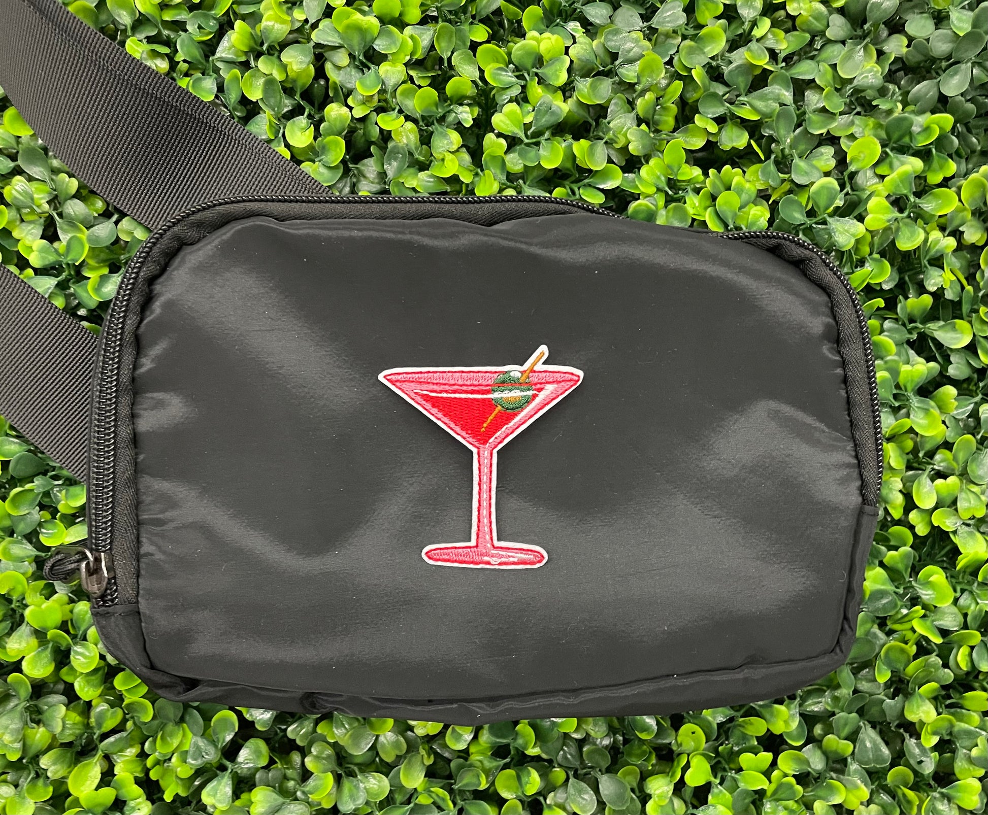 An iron-on patch depicting a red martini glass with a green olive, held onto a black shoulder bag against a green leafy background