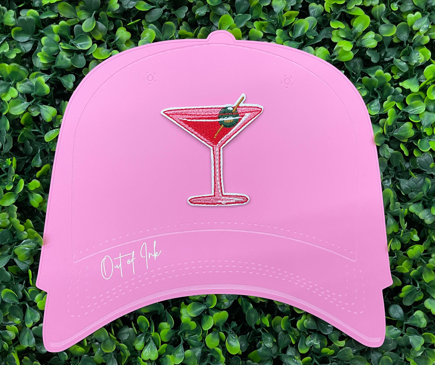 An iron-on patch depicting a red martini glass with a green olive, held onto a pink hat against a leafy background