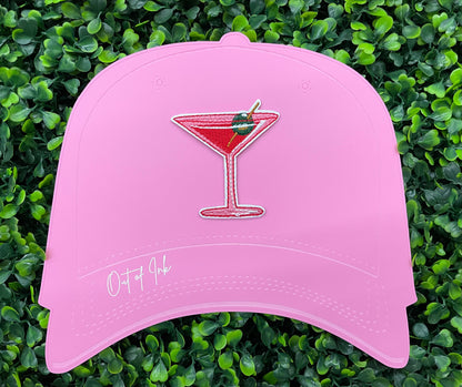 An iron-on patch depicting a red martini glass with a green olive, held onto a pink hat against a leafy background