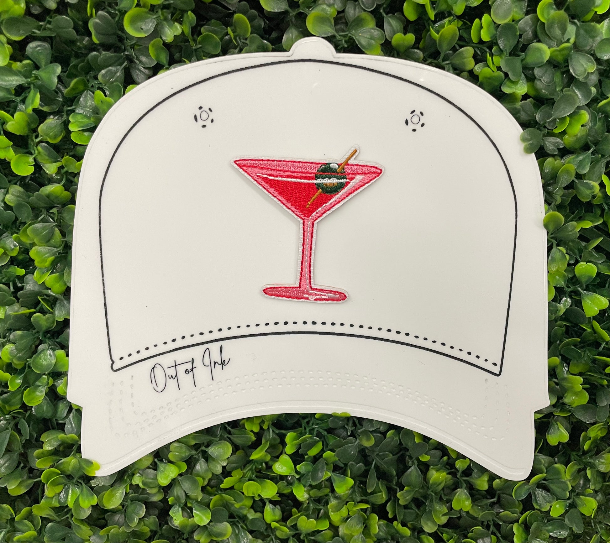 An iron-on patch depicting a red martini glass with a green olive, held onto a white hat against a leafy green background