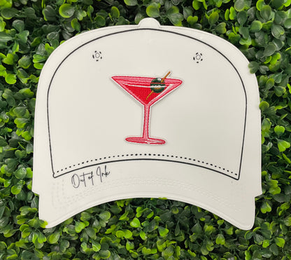 An iron-on patch depicting a red martini glass with a green olive, held onto a white hat against a leafy green background
