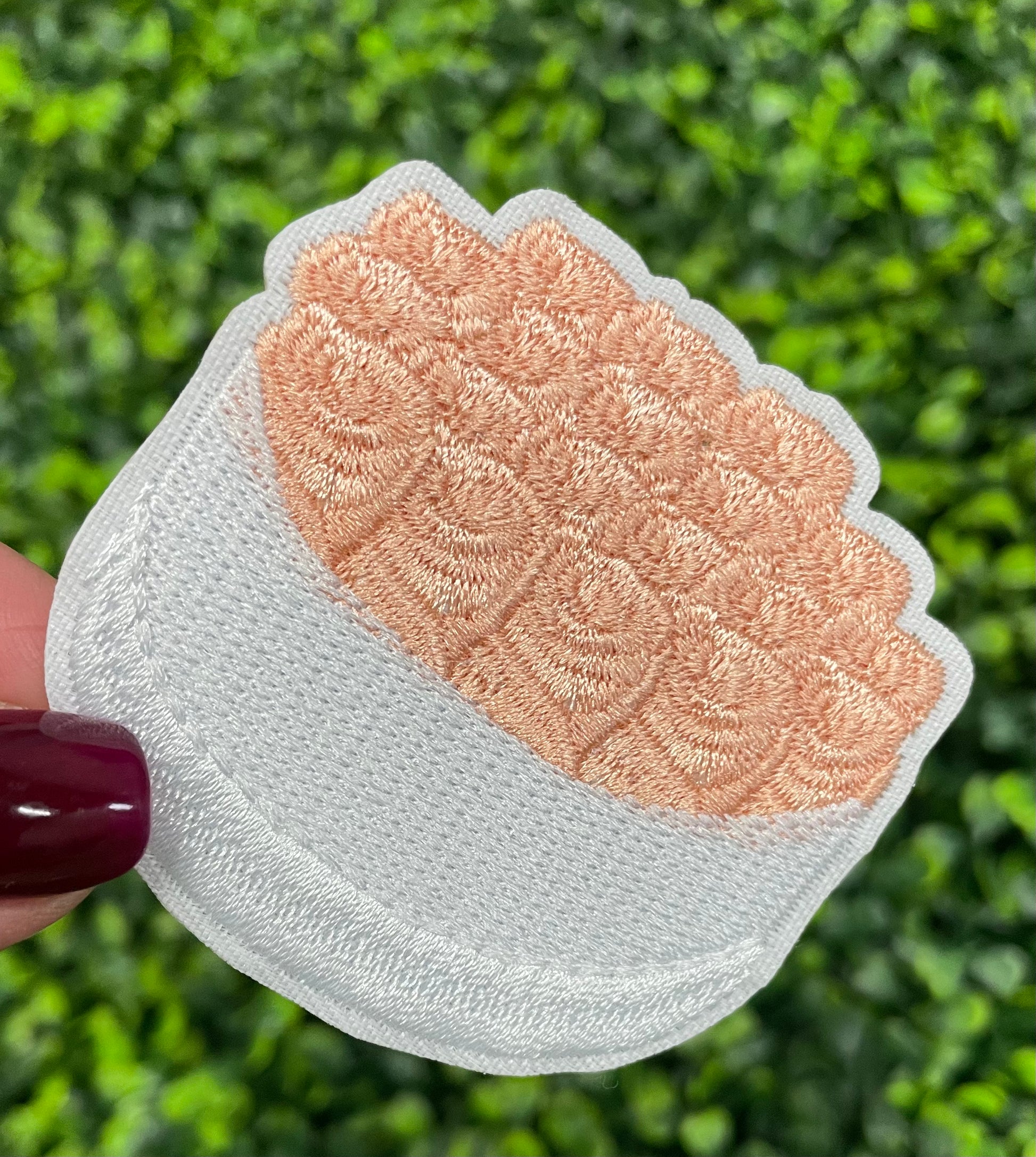 An embroidered patch in the shape of a bundle of roses with pink petals and green leaves, held against a greenery background.