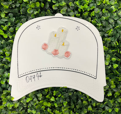 A patch with embroidered candles and roses in pink and yellow colors on a white background, placed on a white hat graphic