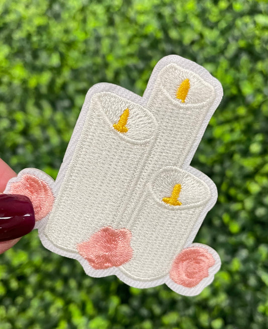 A patch with embroidered candles and roses in pink and yellow colors on a white background, held against a greenery backdrop.