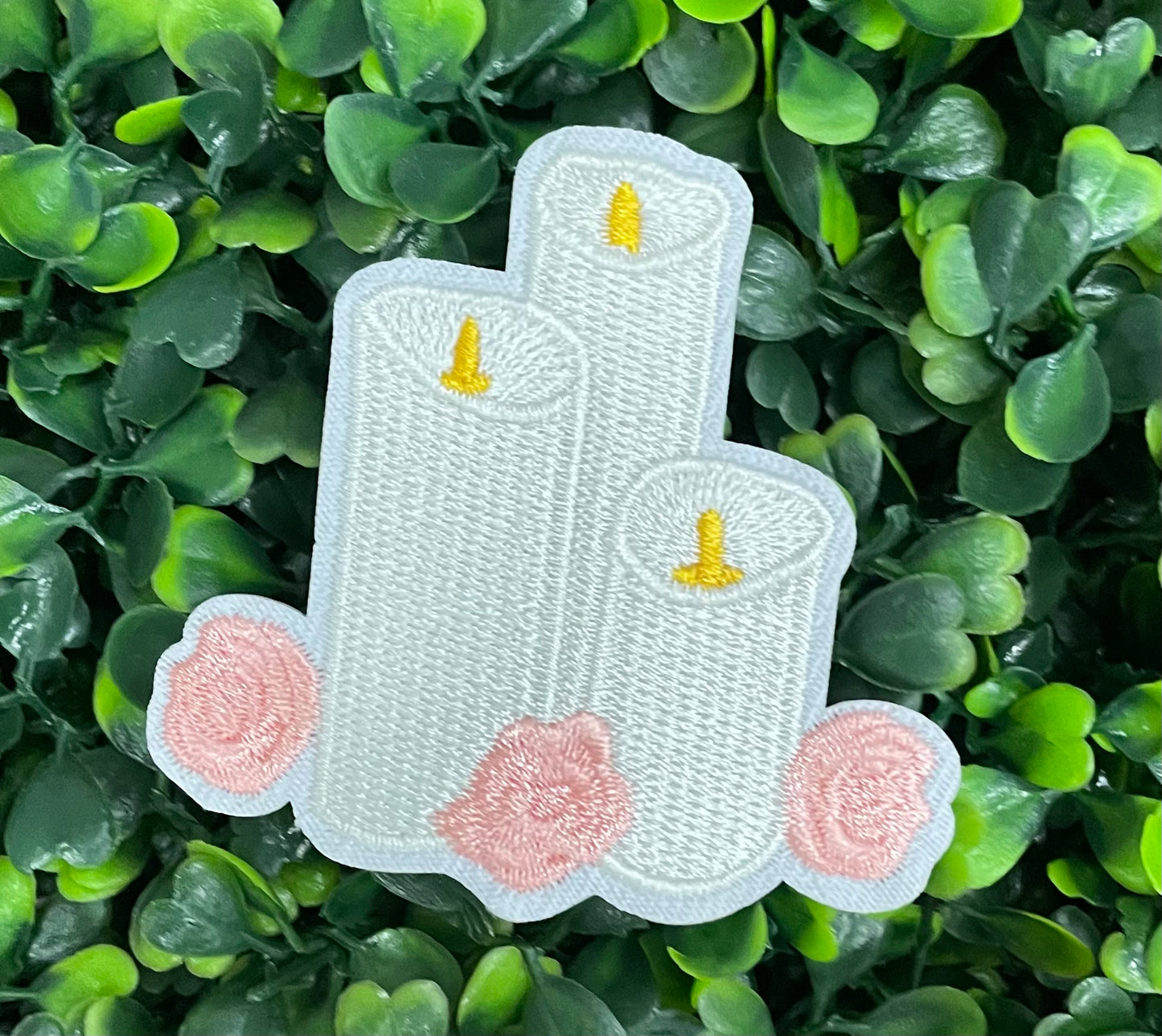 A patch with embroidered candles and roses in pink and yellow colors on a white background, placed on a green leafy background