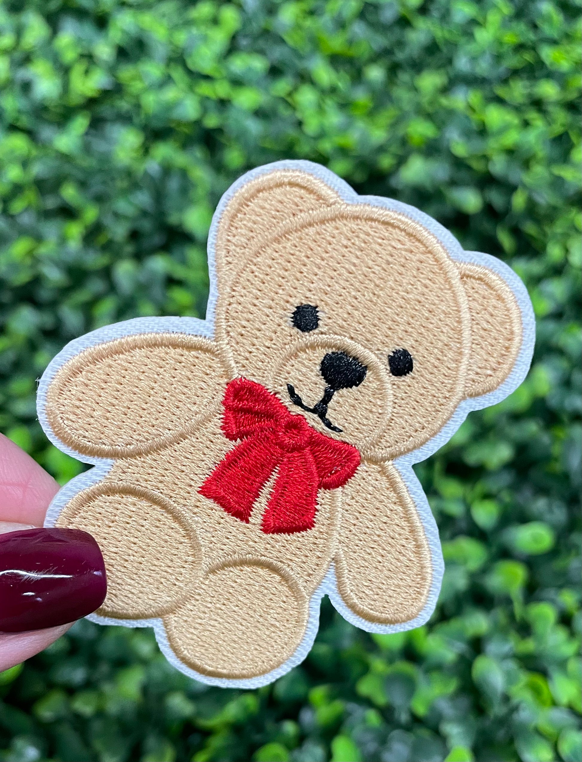 Embroidered Teddy Bear patch with a red bow held by a finger