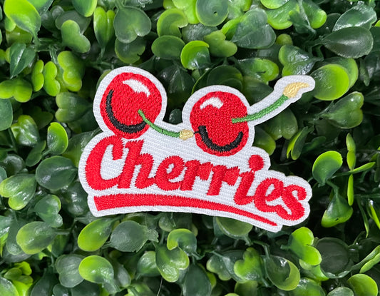 An iron-on patch with an embroidered design of two cherries and the word 'Cherries' below them in red and green colors, placed on a bed of green leaves.