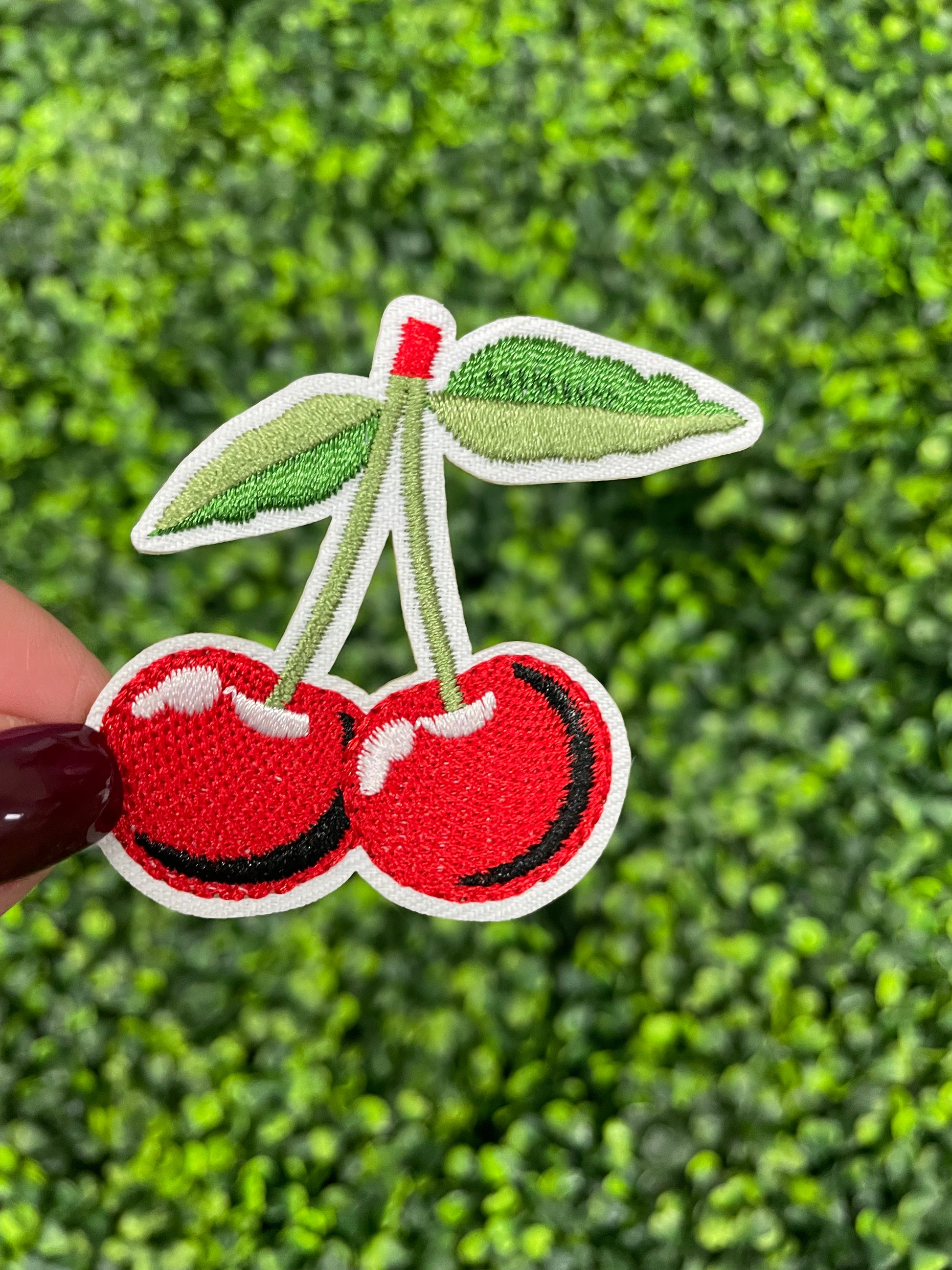 A patch with two red cherries and a green leaf embroidered on it, held against a greenery background.